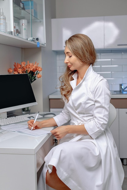 The cosmetologist woman wearing in a white robe is sitting in her office and filling out the documents