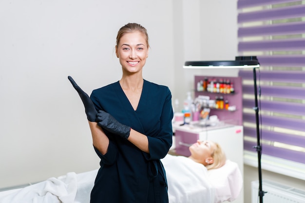 Cosmetology cabinet client lies on couch. Beautician stands puts black gloves on  hand and smiles. Preparation for the procedure of permanent eyebrow makeup.