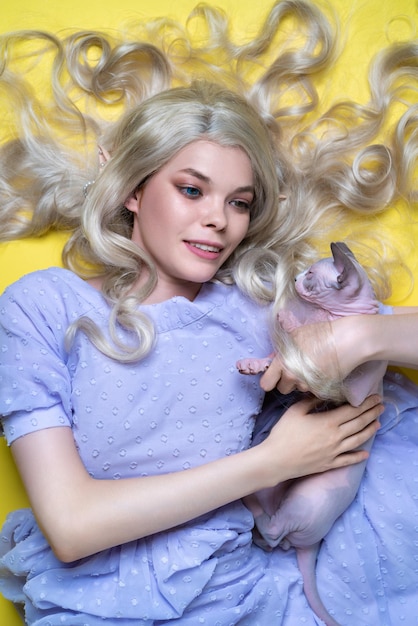 Cosplayer elf young blonde woman in blue dress lies on yellow background hugging sphinx kitten