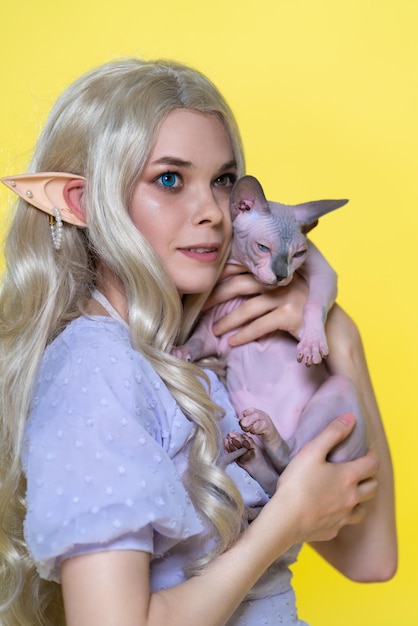 Cosplayer elf young blonde woman in blue dress with curly long hair hugging Sphinx cat to her chest