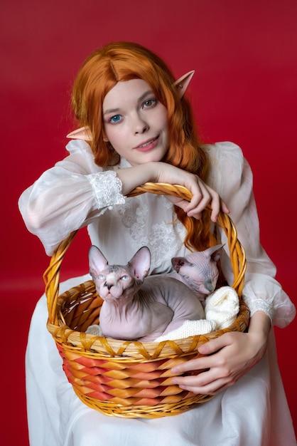 Cosplayer elf young redhead woman smiles leaning on basket in which two Sphinx cats sit calmly