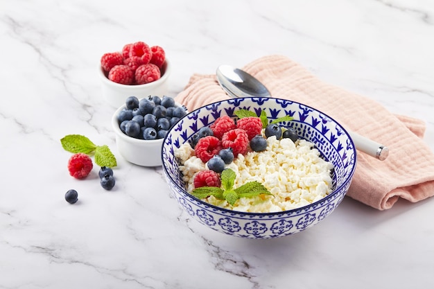 Cottage cheese in bowl decorated with raspberries and blueberries and mint leaves on white