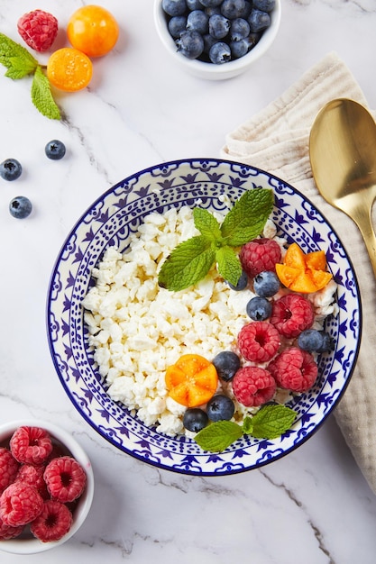 Cottage cheese in bowl garnished with raspberries blueberries physalis and mint leaves on white back