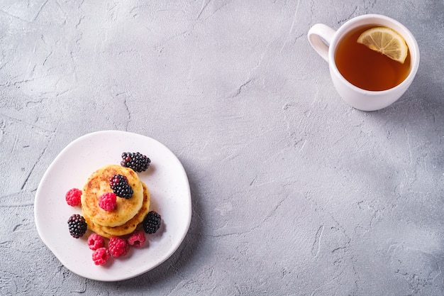 Cottage cheese pancakes, curd fritters dessert with raspberry and blackberry berries in plate near to hot tea cup with lemon slice