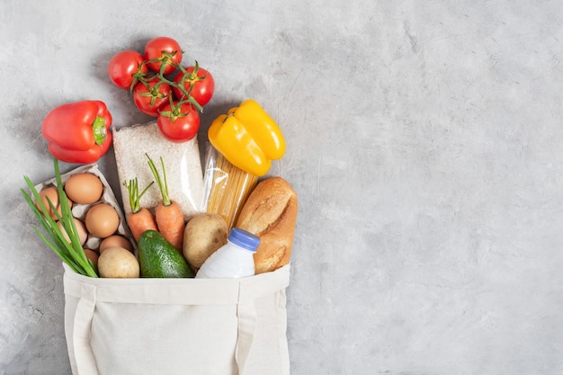 Cotton eco friendly shopping bag full of fresh organic vegetables and grocery products on grey table