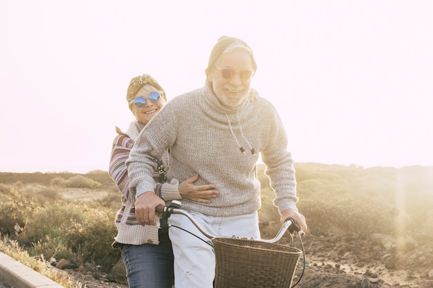 Couple of active cheerful and happy senior retired people enjoy together riding a bike in outdoor leisure activity with sun in backlight - concept of old age and fun joyful with no limit