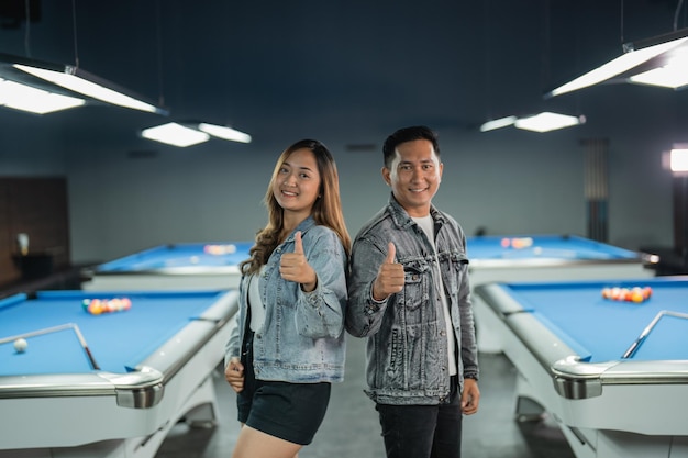 Couple of asian billiard players standing back to back with thumb up with the billiard tables at the