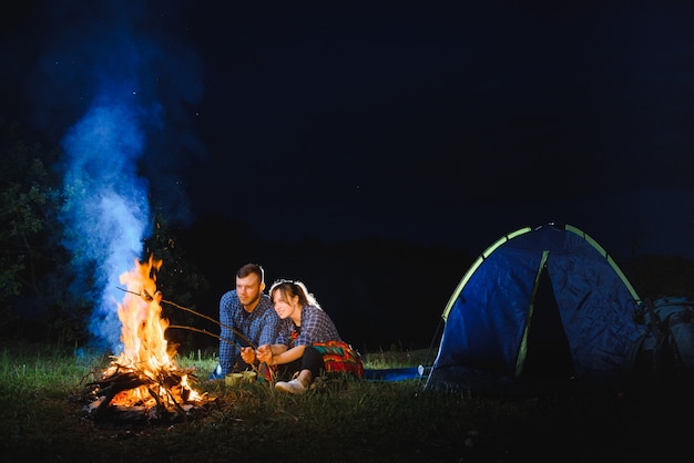 Couple baking sausages on the fire, and relaxing by the campfire in the forest in the night