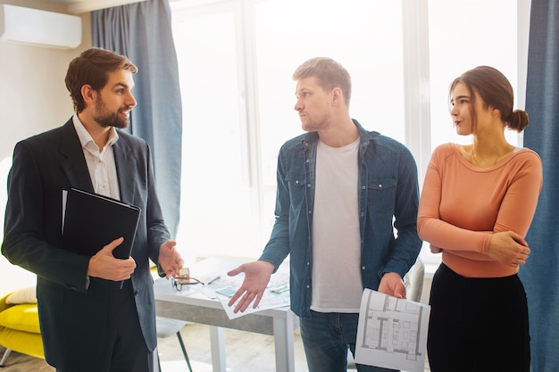 Couple buy or rent apartment together. They look confused at realtor. Cliets have doubts. Serious realtor look at them. People in one room