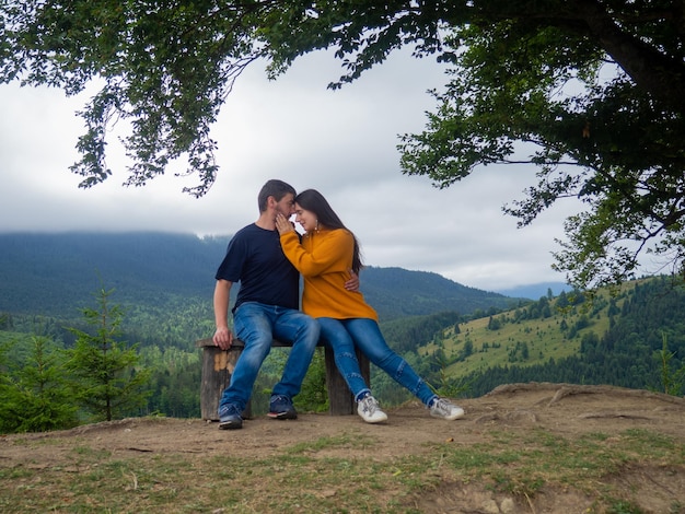 Couple in casual clothes sit under large tree with forest background