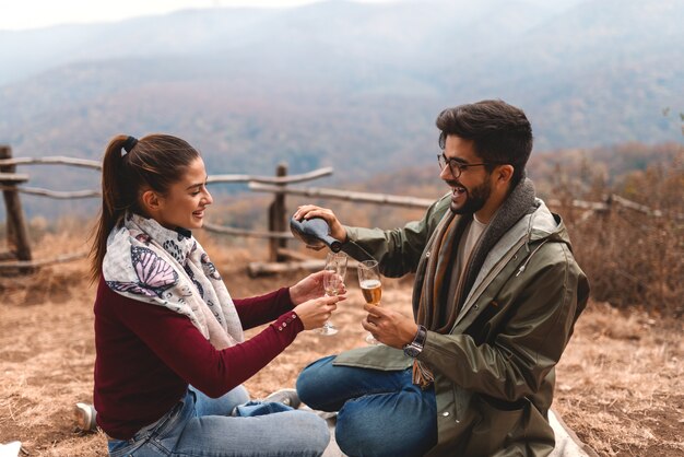 Couple celebrating anniversary at picnic. Man pouring champagne into glasses. Autumn time.