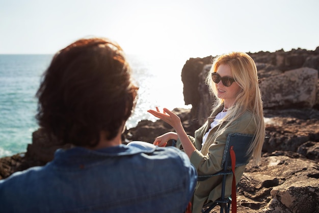 Couple chatting and relaxing in camping chairs on beach rocks near ocean