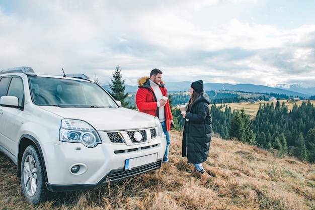 Couple drinking coffee to go at the top of the hill near car with beautiful view of mountains. car travel concept