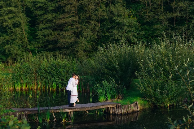 Couple in embroidered clothes hugging on a wooden pier of a lake trees and reeds by the lake