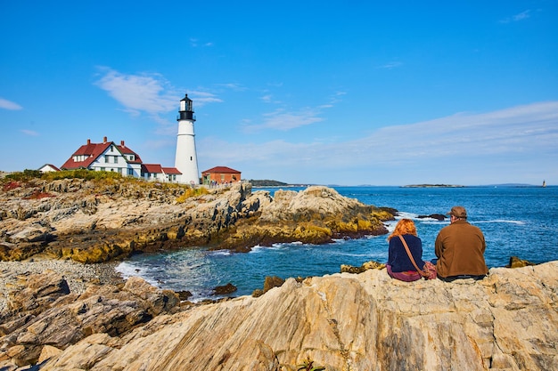 Couple enjoys view of lighthouse on Maine coastline sitting on rocks