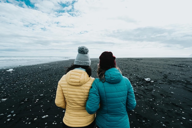 Couple female in yellow rain clothes in the black sand diamond beach near Jokulsarlon during a road trip travel exploring wild lands of IcelandAdventure vacations healthy lifestyle backpacking