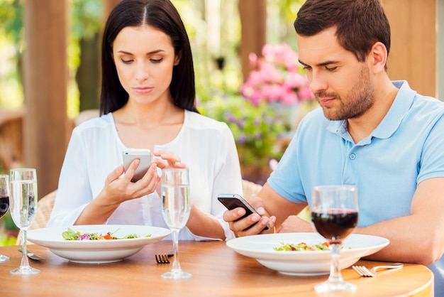 Couple from digital age. Young couple typing something on their smart phones while relaxing in outdoors restaurant together