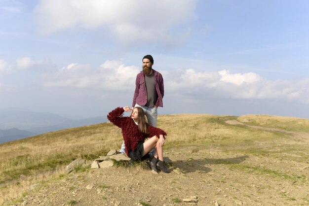 Couple of handsome bearded hipster in checkered shirt and pretty cute sexy young fashion girl or woman sitting on stone with ax on mountain top on natural background