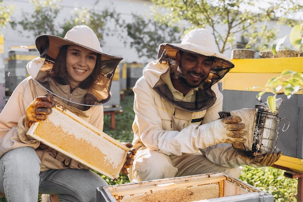 Photo couple of happy smiling beekeepers working with beekeeping tools near beehive at bee farm