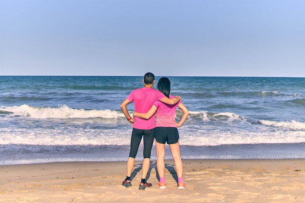 Couple holding each other in front of the sea after running training in the morning. Wearing pink sportswear. Summer day at the beach.