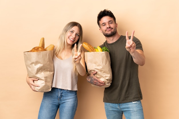 Couple holding grocery shopping bags over smiling and showing victory sign