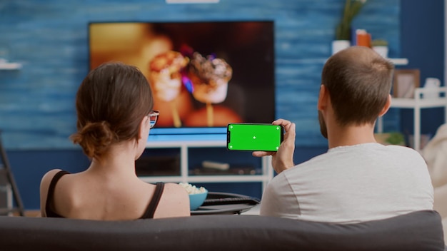 Couple at home watching online tutorials on green screen smartphone while enjoying free time sitting on couch. Man and woman looking at webinar or social media videos on chroma key mobile phone.