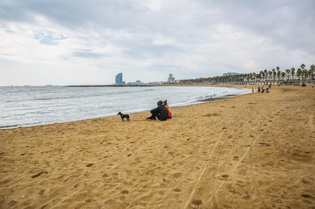 Couple is sitting at the autumn beach Dog is walking on the sand Atmosphere photo Barceloneta Man and woman in love