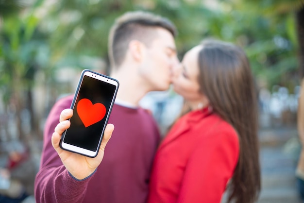 Couple kissing while showing a smartphone with an heart shape in it dating application concept