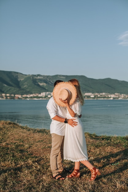 A couple in love after their engagement embrace on the beach in the sunset. Kiss the newlyweds, covered with a hat. A girl in a white dress with a guy in a romantic place