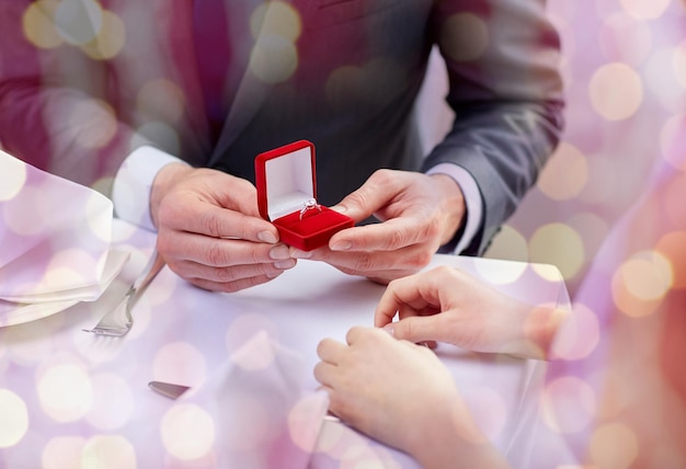 couple, love, engagement and holiday concept - close up of excited young woman and boyfriend giving her ring at restaurant over pink holidays lights background