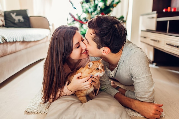 Couple in love lying by Christmas tree and playing with cat at home. Happy man and woman kissing