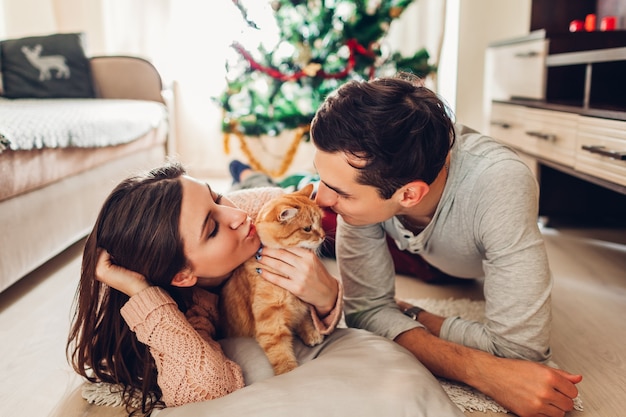 Couple in love lying by Christmas tree and playing with cat at home