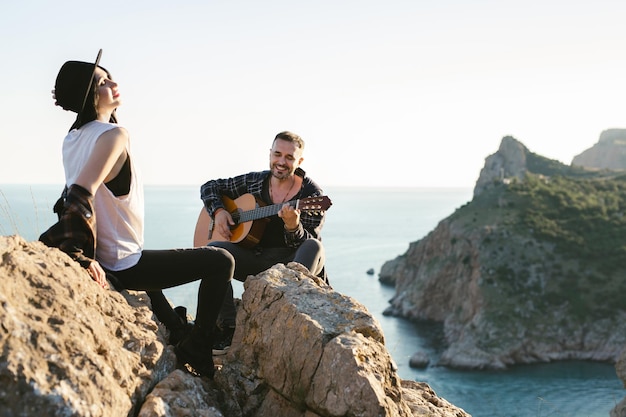 Couple in love on a mountaintop overlooking the sea Man plays the guitar a woman in a hat sings