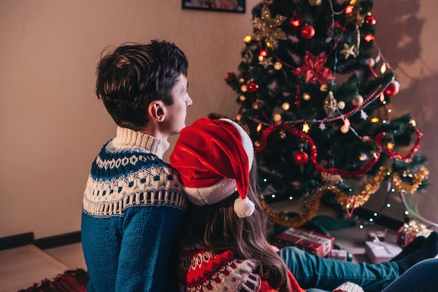 Couple in love sitting next to a Christmas tree, wearing Santa's hat and hugging.