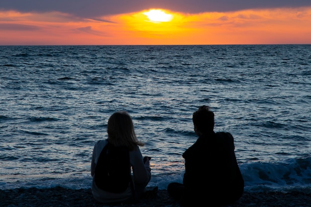 Couple in love watching sunset together on beach travel summer holidays. People silhouette from behind sitting enjoying view sunset sea on tropical destination vacation. Romantic couple on the beach