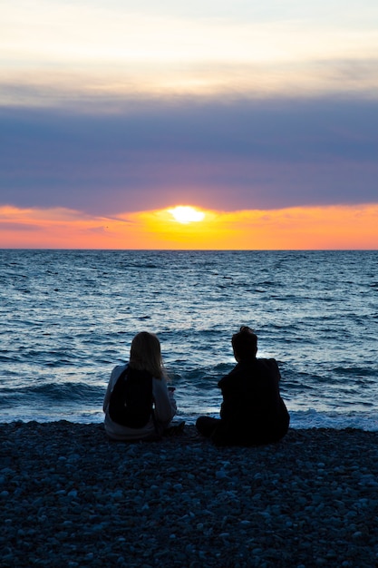 Couple in love watching sunset together on beach travel summer holidays. People silhouette from behind sitting enjoying view sunset sea on tropical destination vacation. Romantic couple on the beach