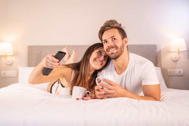 A couple in love watching TV on top of the hotel bed in pajamas, lifestyle of a loving couple.