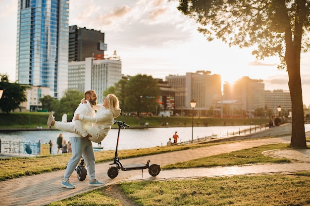 A couple in love in white clothes stands in the city at sunset near an electric scooter.