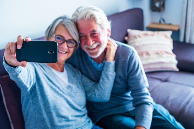 Couple of mature people having fun at home sitting on the sofa taking a selfie together smiling and looking at the phone - two retired pensioners enjoying the technology indoors