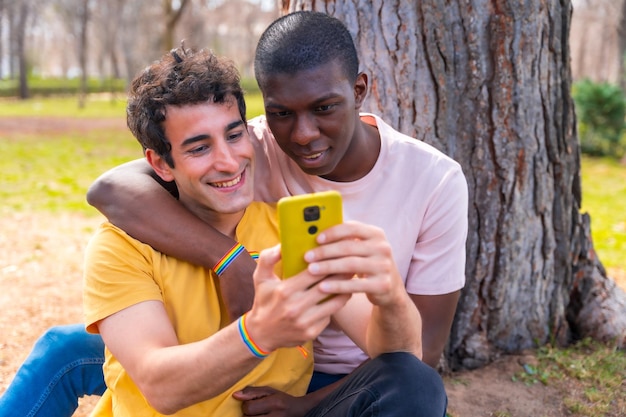 Couple of multiethnic men in a park lgbt concept sitting next to a tree having fun with a yellow mobile