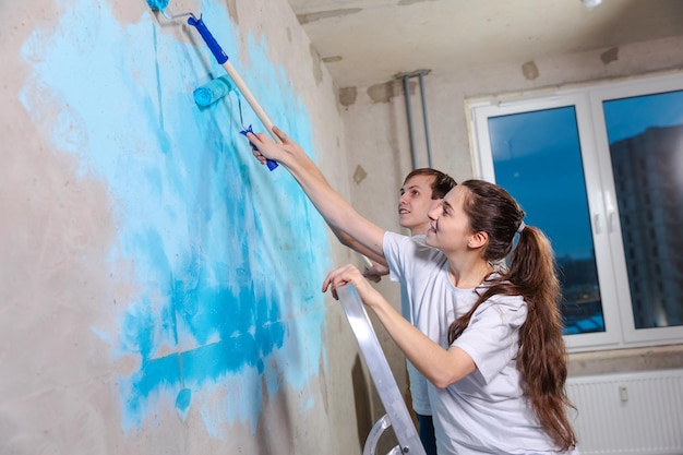 Couple in new home during repair works painting wall together Happy family holding paint roller painting wall with blue color paint in new house Home renovation DIY renew home concept