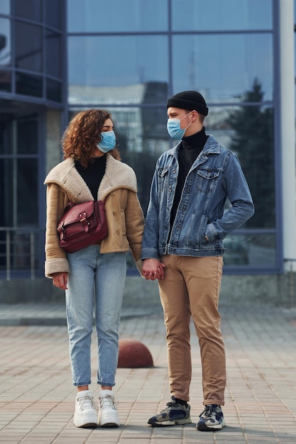 Couple in protective masks have a walk outdoors in the city near business building at quarantine time Conception of coronavirus