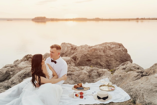 A couple sits on a blanket in front of a lake and the sunset is visible.