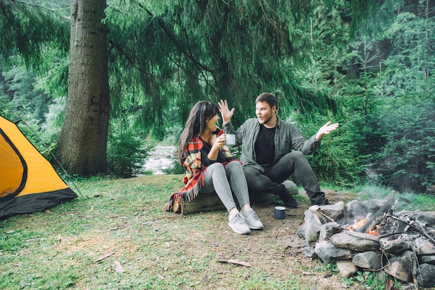 Couple sitting near camp fire and drinking tea and telling stories . tent and suv on background. union with nature