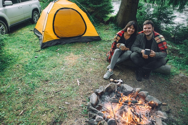 Couple sitting near camp fire and drinking tea and telling stories tent and suv on background