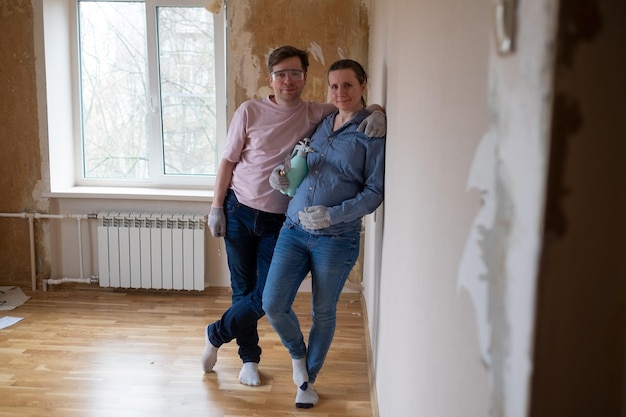 Couple smiling at camera standing near wall redecorating their new flat