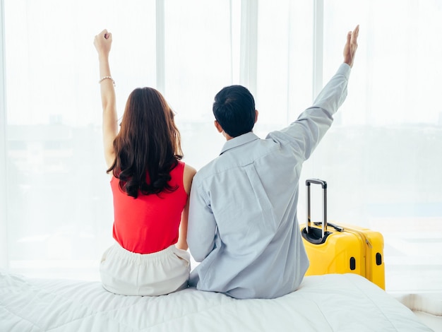 Couple of tourists happy holiday Summer vacation Portrait of back view of young Asian man and woman raising hand with happy on white bed with suitcase near curtain at the window in hotel room