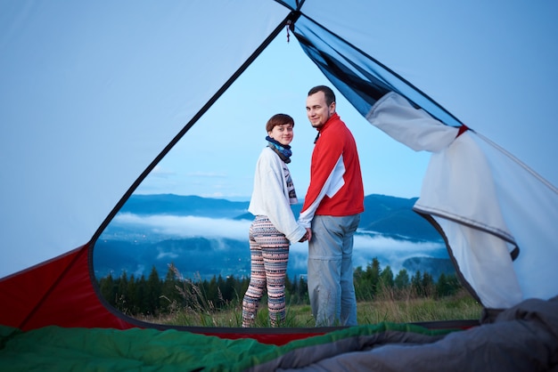 Couple of tourists holding hands looking at the camera against the backdrop of the mighty mountains in the morning fog at dawn. View from inside a tent