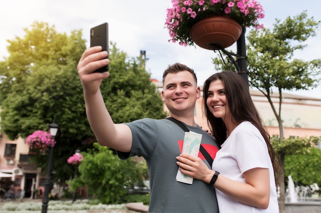 A couple of tourists in love with map in hands doing a selfie. Girlfriend and boyfriend are looking into camera and smiling for a good photo shot. Handsome heterosexual couple are walking the street.