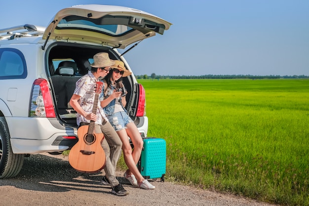 Couple of traveler sitting on hatchback of car and looking at the picture on camera 